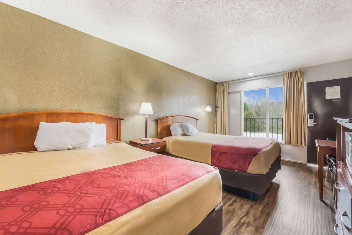 A hotel room with two beds, a desk, a chair, a lamp, and a view of the outdoors.