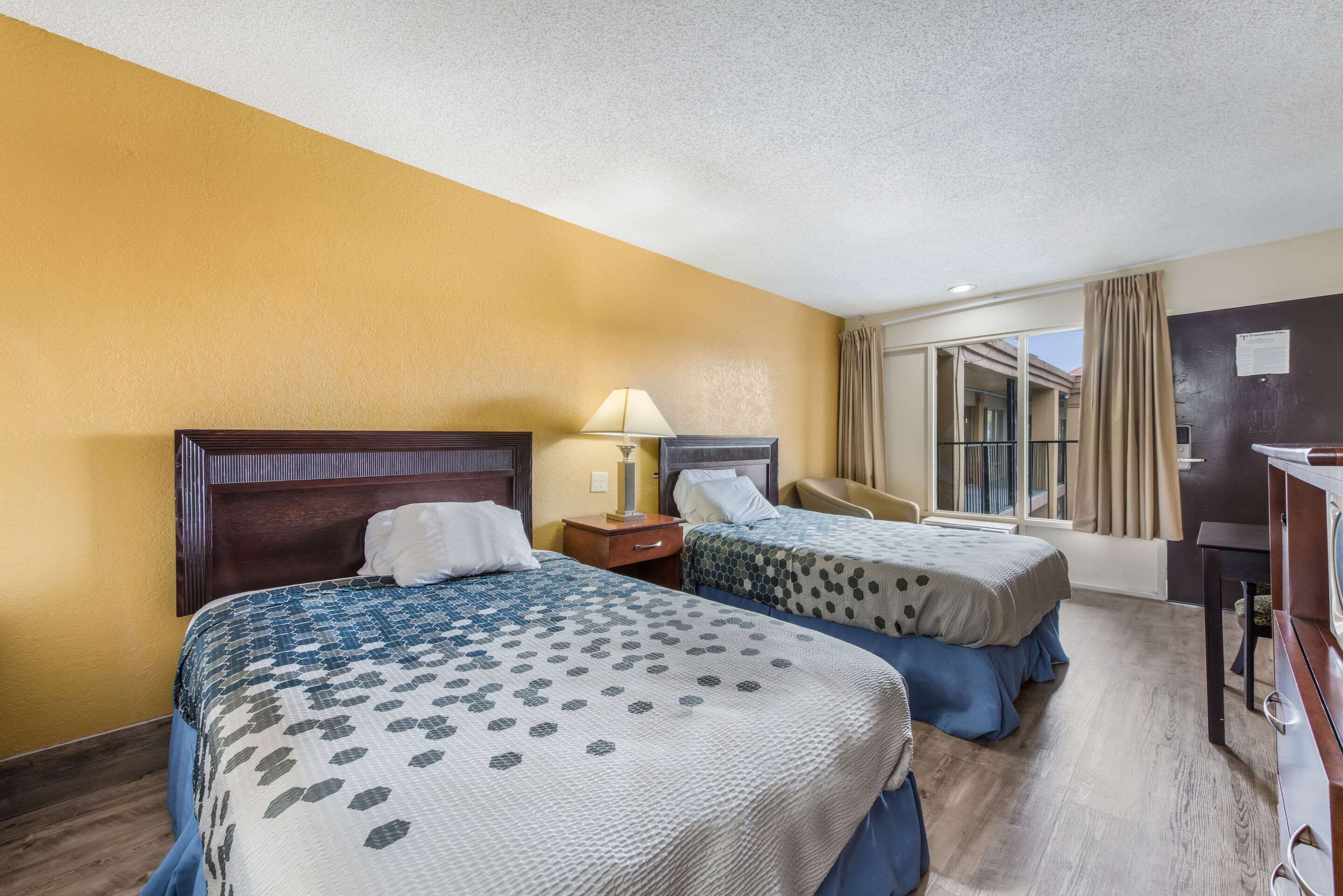 Habitación de hotel con dos camas, un escritorio, una silla y un ventanal con cortinas.