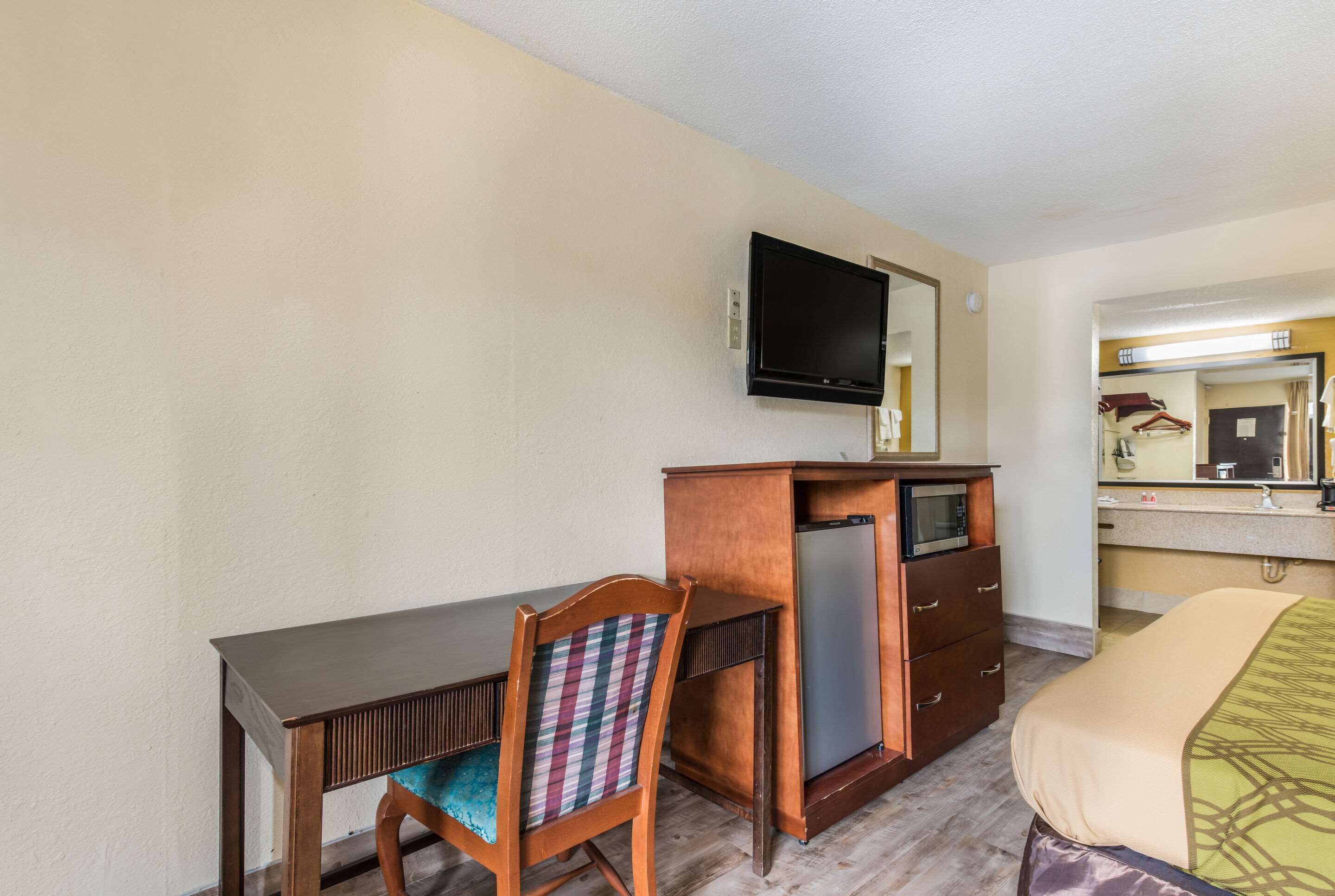 Une chambre d’hôtel avec un bureau en bois, une chaise dotée d’un coussin à carreaux, un téléviseur fixé au mur, un minibar, un lit avec une couvre-lit à motifs et une salle de bain avec un miroir et un lavabo.