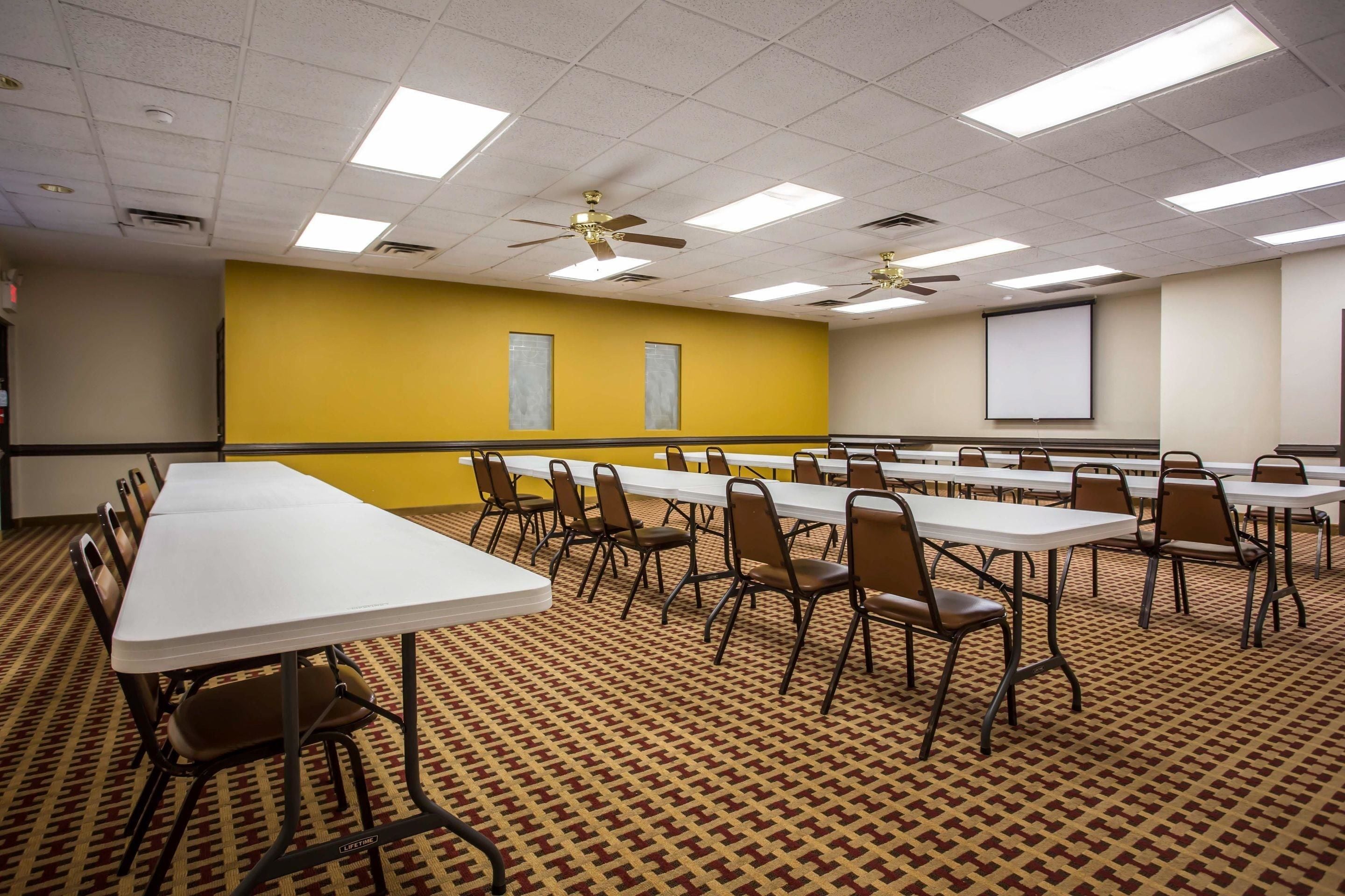 A conference room with long tables and chairs, ceiling fans, and a projector screen on the wall.