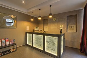 A reception area with a modern design, featuring a reception desk with illuminated glass panels displaying text, pendant lighting, a large mirror, a clock, and a framed certificate on the wall.