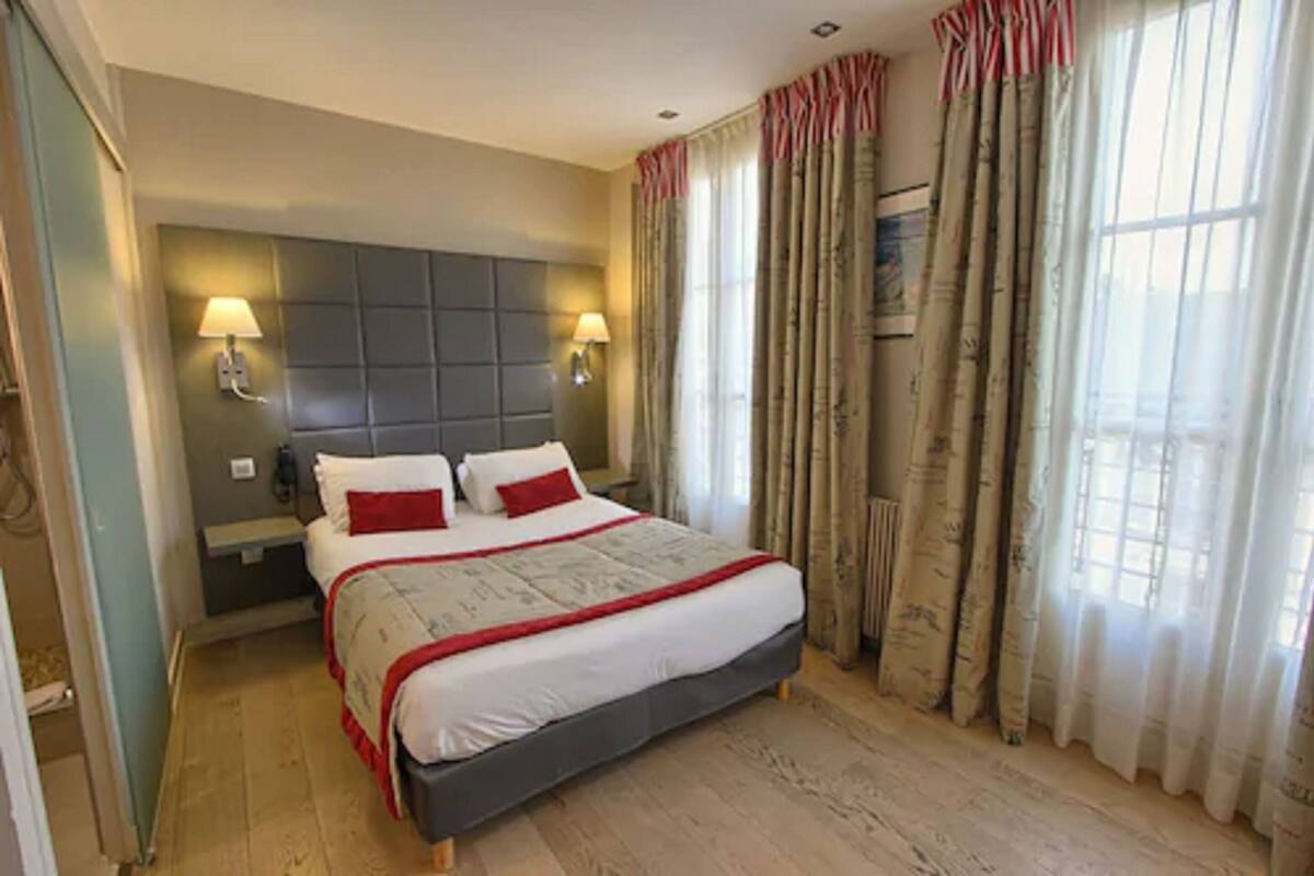 A bedroom with a large bed, two bedside lamps, a headboard, and a view of the outdoors through curtains.