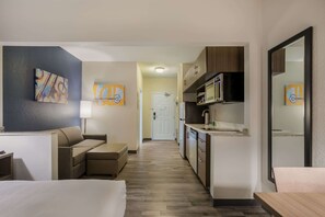 A hotel room with a bed, a sofa, a kitchenette, and artwork on the walls.