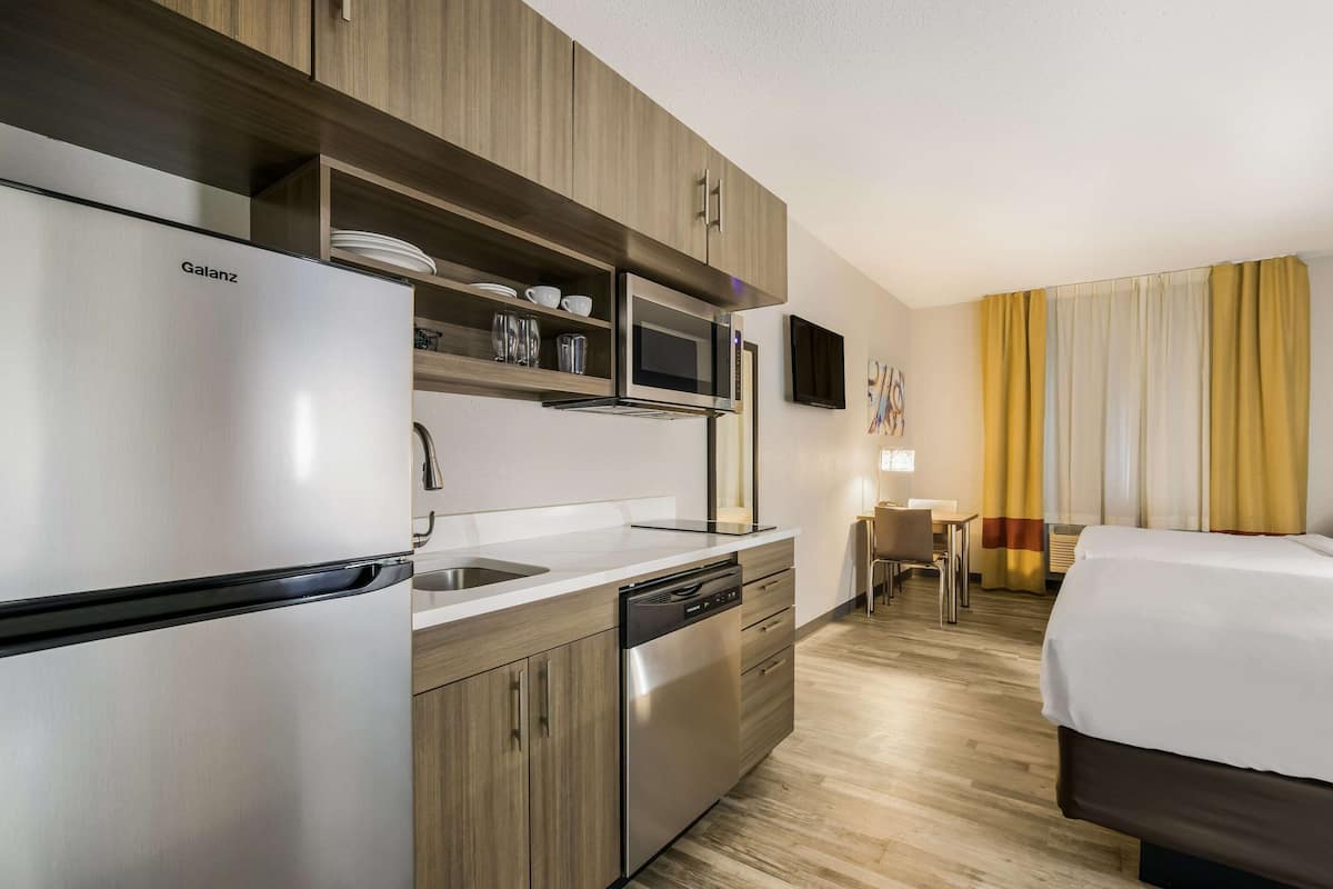 Camera d'albergo compatta con angolo cottura, che include un frigorifero Galanz, un forno a microonde e una lavastoviglie. È presente una piccola zona pranzo con tavolo e sedie, e un letto con copripiumino bianco.