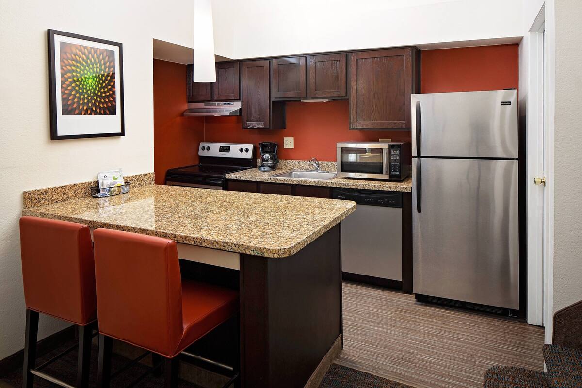 A compact kitchen with granite countertops, a stainless steel refrigerator, a microwave, and a coffee maker. There are two red bar stools and a framed picture on the wall.