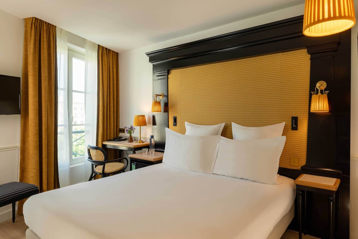 A hotel room with a large bed, a desk with a lamp, a chair, and a TV on the wall.