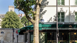 A street view of a building with a green awning, outdoor seating, and a clock tower in the background. - Hôtel Observatoire Luxembourg (Paris)