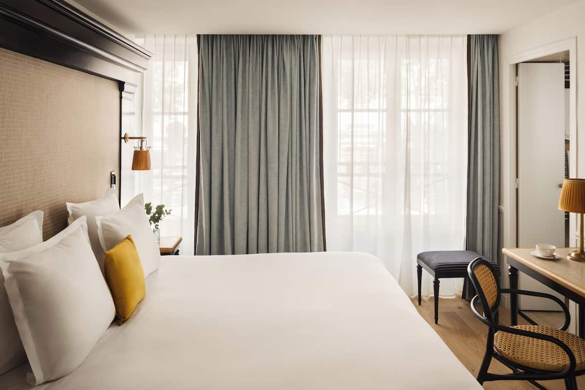 Une chambre d’hôtel avec un grand lit, un bureau avec une chaise, une lampe et une vue à travers des rideaux transparents.