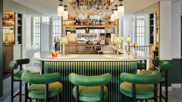 A bar area with a central bar counter, green upholstered bar stools, and a variety of bottles and glasses on shelves. - Hôtel Observatoire Luxembourg (Paris)