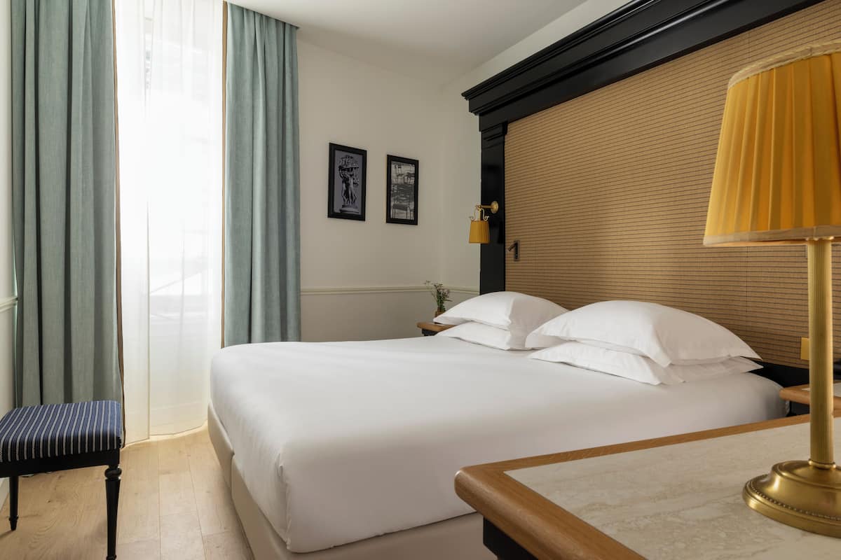 A hotel room with a large bed, a bench, a bedside table with a lamp, and two framed pictures on the wall.
