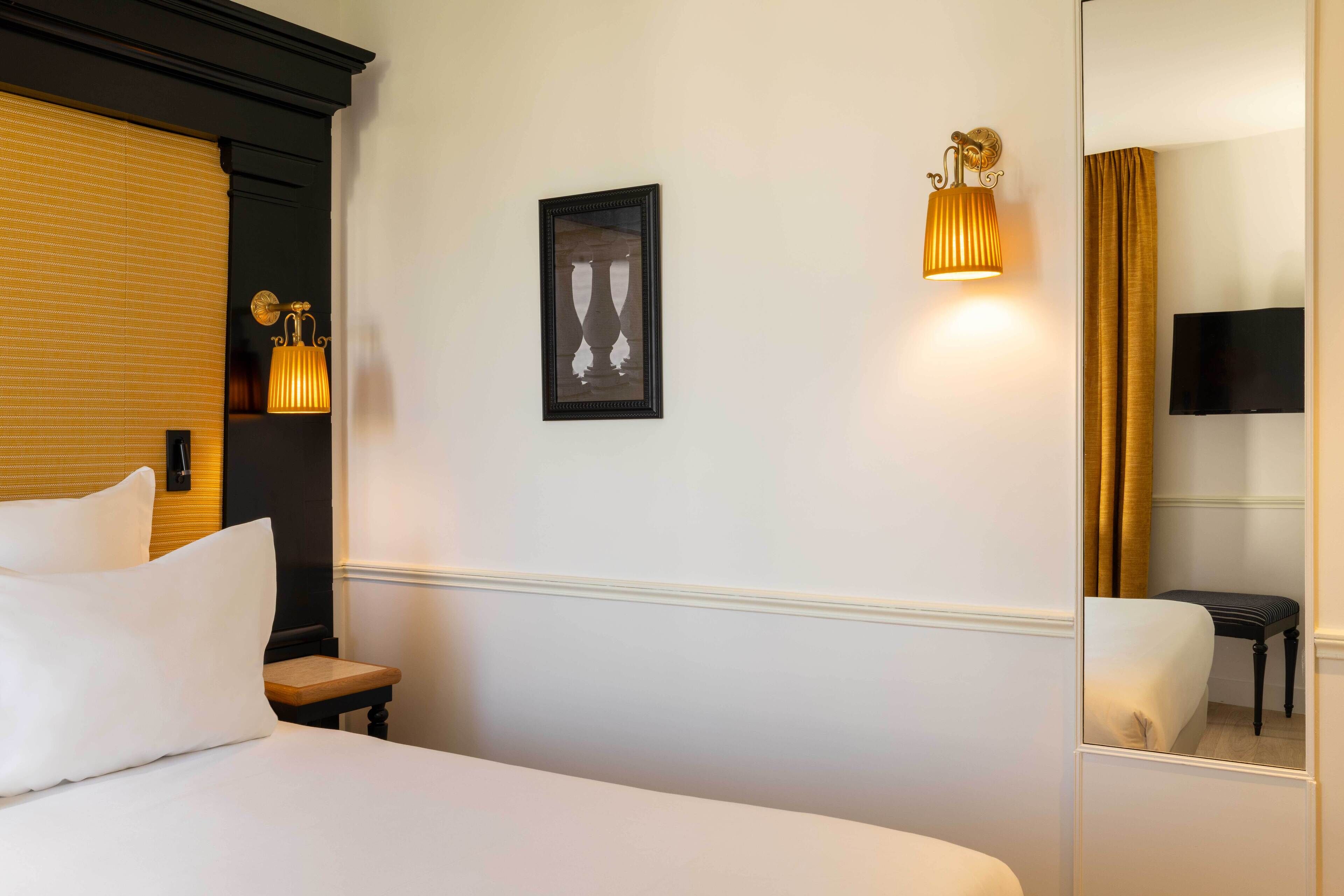 A hotel room with a bed, a nightstand, a wall-mounted lamp, a framed picture, a mirror, and a television.
