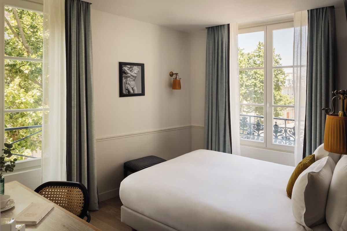 A hotel room with a bed, a desk with a chair, a small stool, a framed picture on the wall, and a window with curtains.