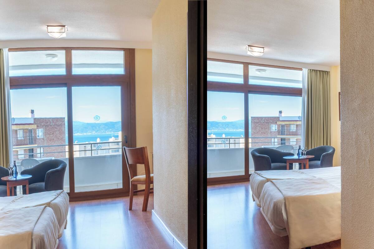 A hotel room with a bed, two chairs, a small table, and a view of the sea.