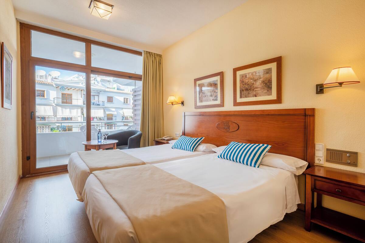 A hotel room with two beds, a desk, a chair, a balcony with a view of buildings, and a wall-mounted lamp.