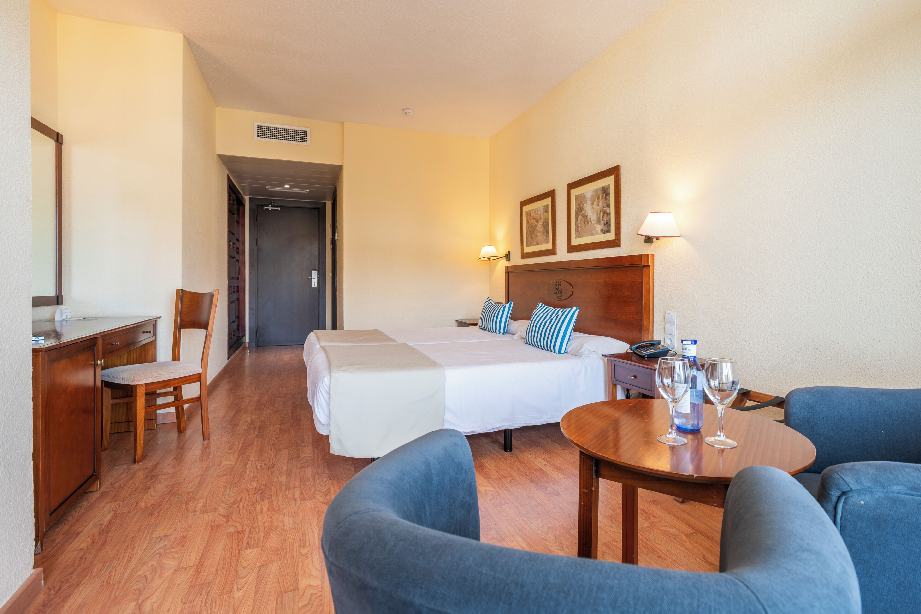 A hotel room with a bed, two bedside tables, a desk with a chair, a round table with two chairs, and a door leading to another room.