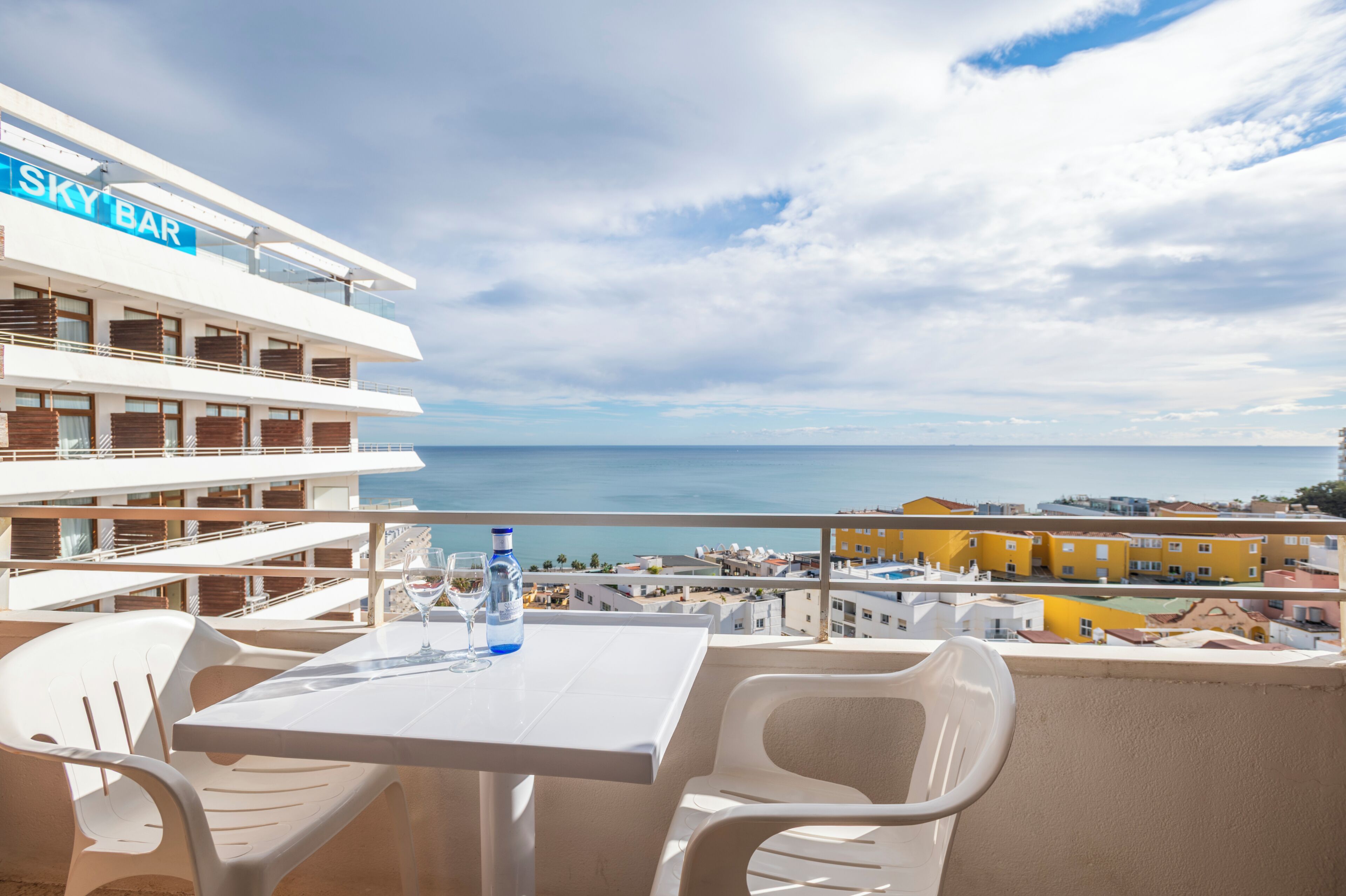 A balcony with a table and chairs, overlooking a coastal view with buildings in the background.