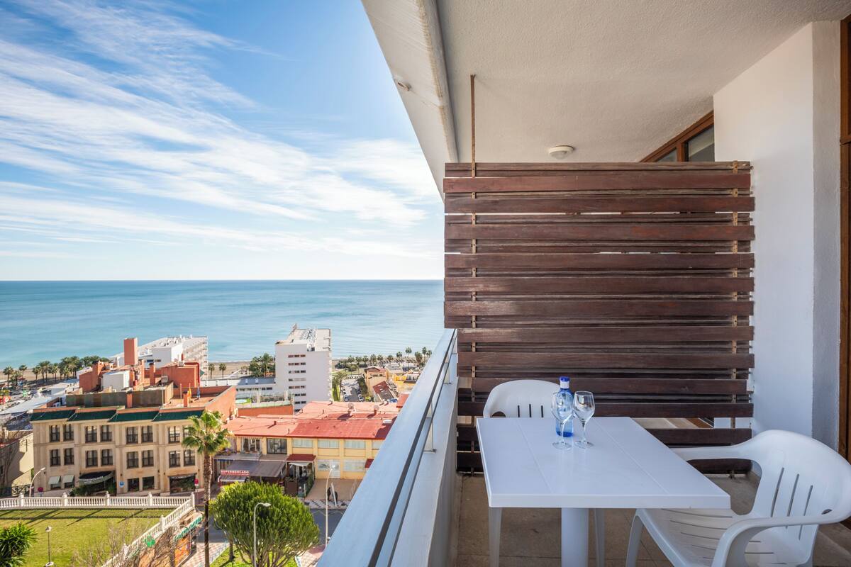 A balcony with a white table and chairs, overlooking a cityscape and the sea.