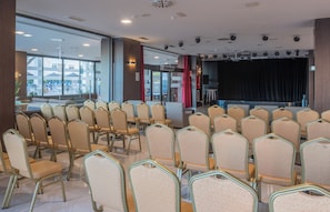 Une salle de conférence avec des rangées de chaises, une scène avec des rideaux et de grandes fenêtres donnant sur un espace extérieur aménagé avec des sièges.