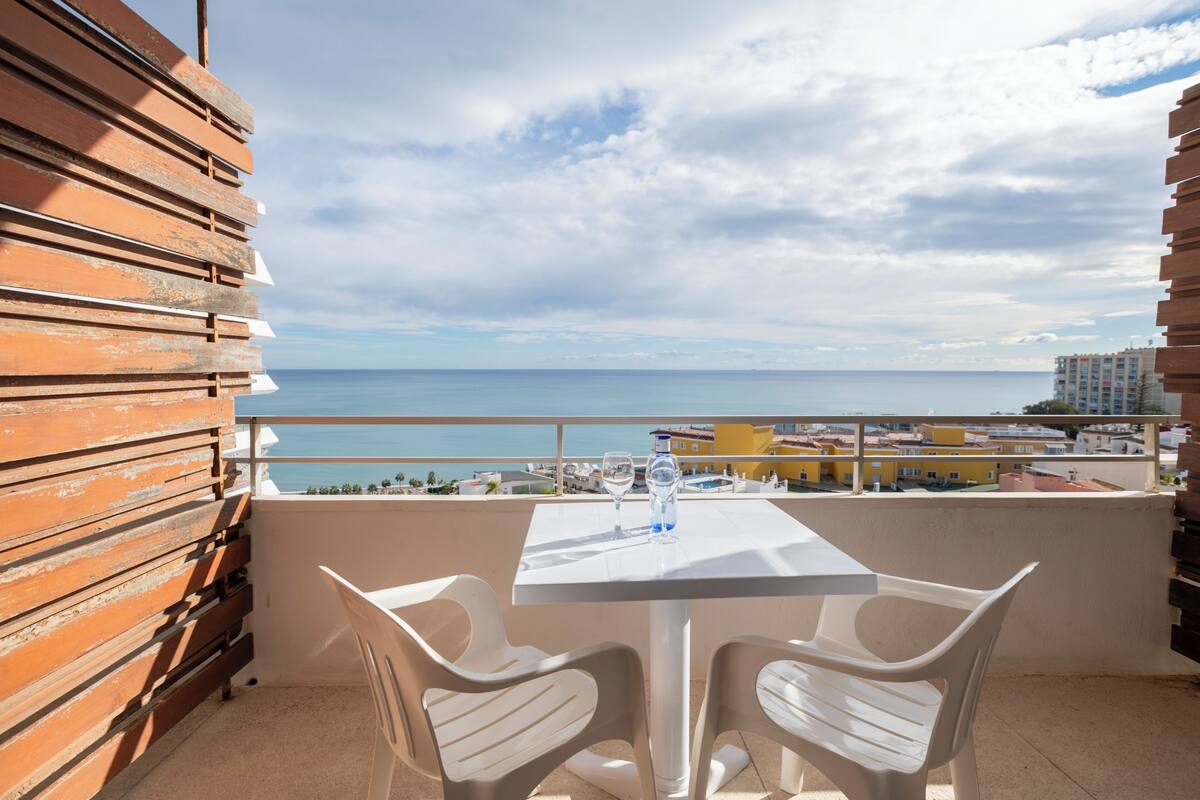 A balcony with a table and chairs, overlooking a coastal view with buildings in the distance.