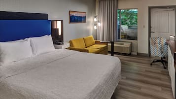 A hotel room with a large bed, a yellow armchair, a desk with a chair, a window with curtains, and a door.