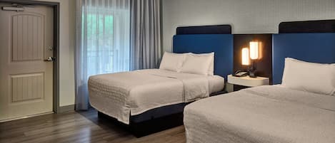 A hotel room with two beds, a door, a window with curtains, a nightstand with a lamp, and a patterned wall.