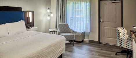 Habitación de hotel con cama, una silla, escritorio, lámpara, calefactor y puerta.