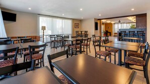 Une salle à manger d’hôtel avec plusieurs tables et chaises, un téléviseur fixé au mur et une grande fenêtre avec des rideaux.
