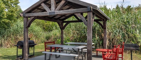 Un pabellón de madera con mesas de picnic, bancos, una parrilla y un banco rojo.