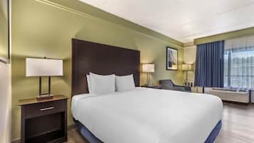 Ein Hotelzimmer mit einem großen Bett, Nachttischen, einem Sessel, einem Schreibtisch, einem Fenster mit Vorhängen und einer Wand-Klimaanlage.