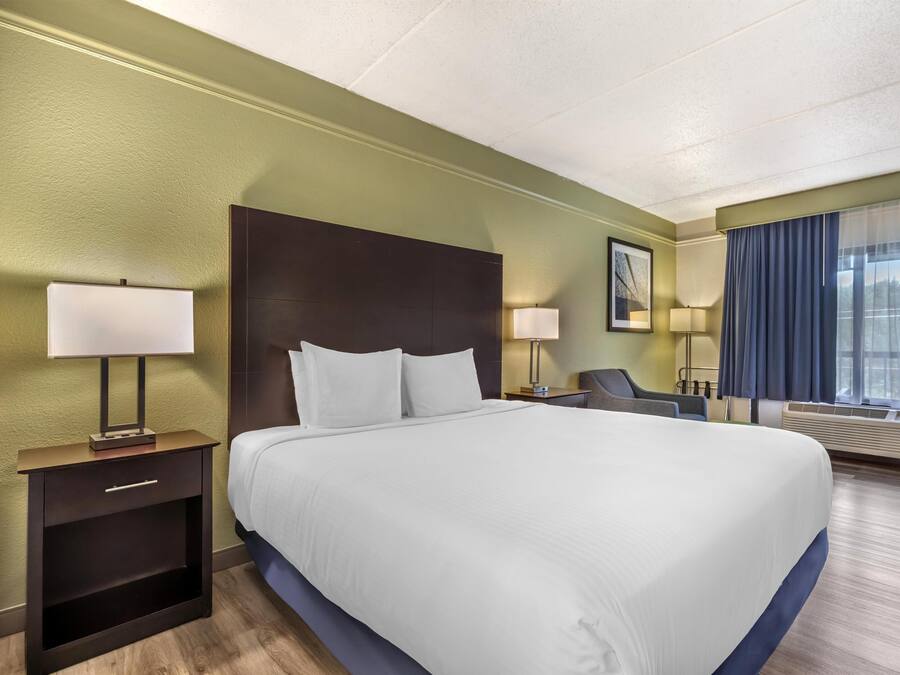 Ein Hotelzimmer mit einem großen Bett, Nachttischen, einem Sessel, einem Schreibtisch, einem Fenster mit Vorhängen und einer Wand-Klimaanlage.