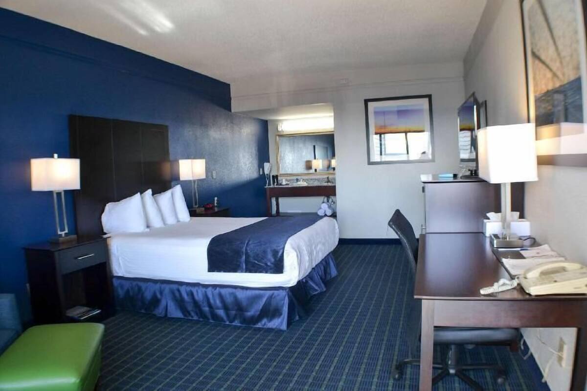 A hotel room with a bed, desk, chair, and a small green stool.