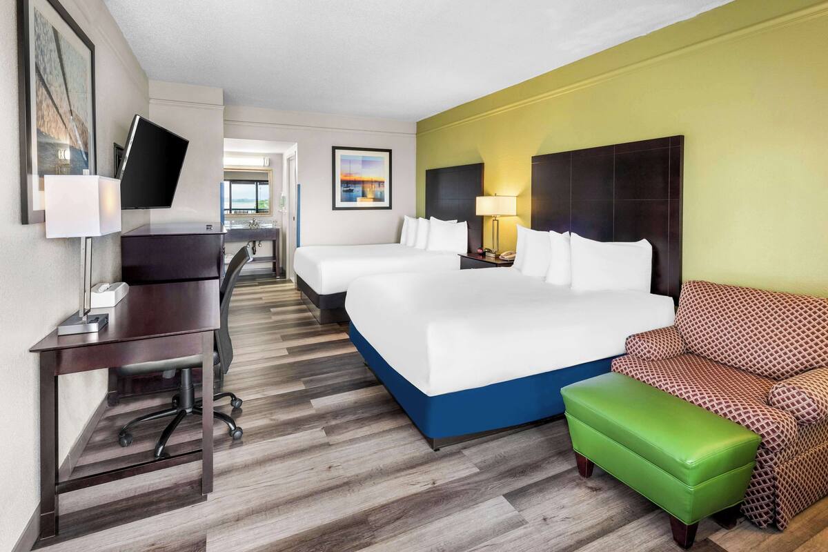 A hotel room with a large bed, a desk with a computer, a chair, a green ottoman, and a sofa.