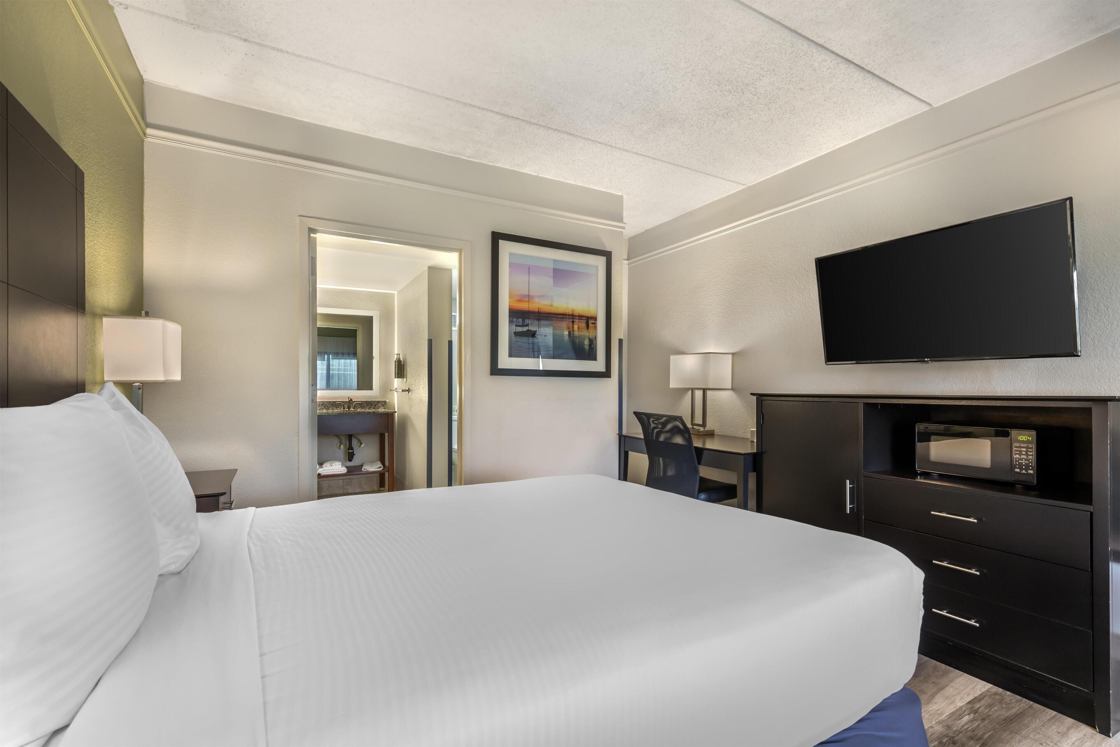 A hotel room with a large bed, a flat-screen TV, a desk with a chair, and a bathroom visible through an open door.