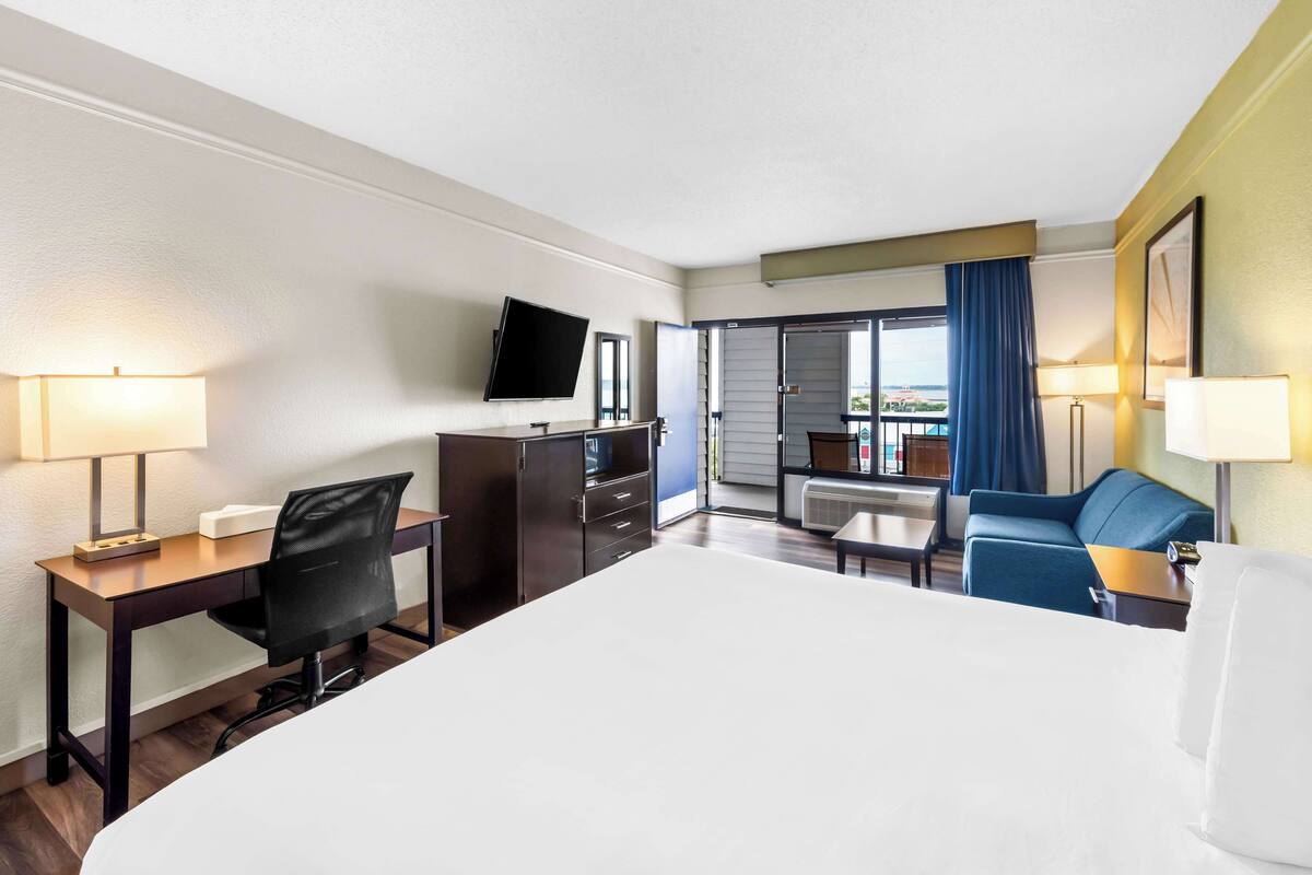 A hotel room with a large bed, a desk with a chair, a TV, a balcony with a view, and a blue sofa.