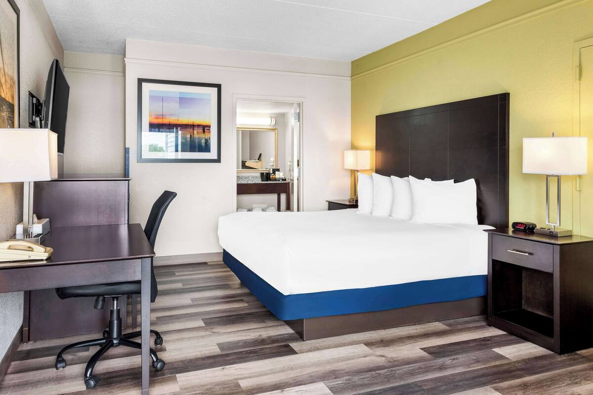 A hotel room with a large bed, a desk with a chair, a nightstand with a lamp, and a bathroom visible through an open door.