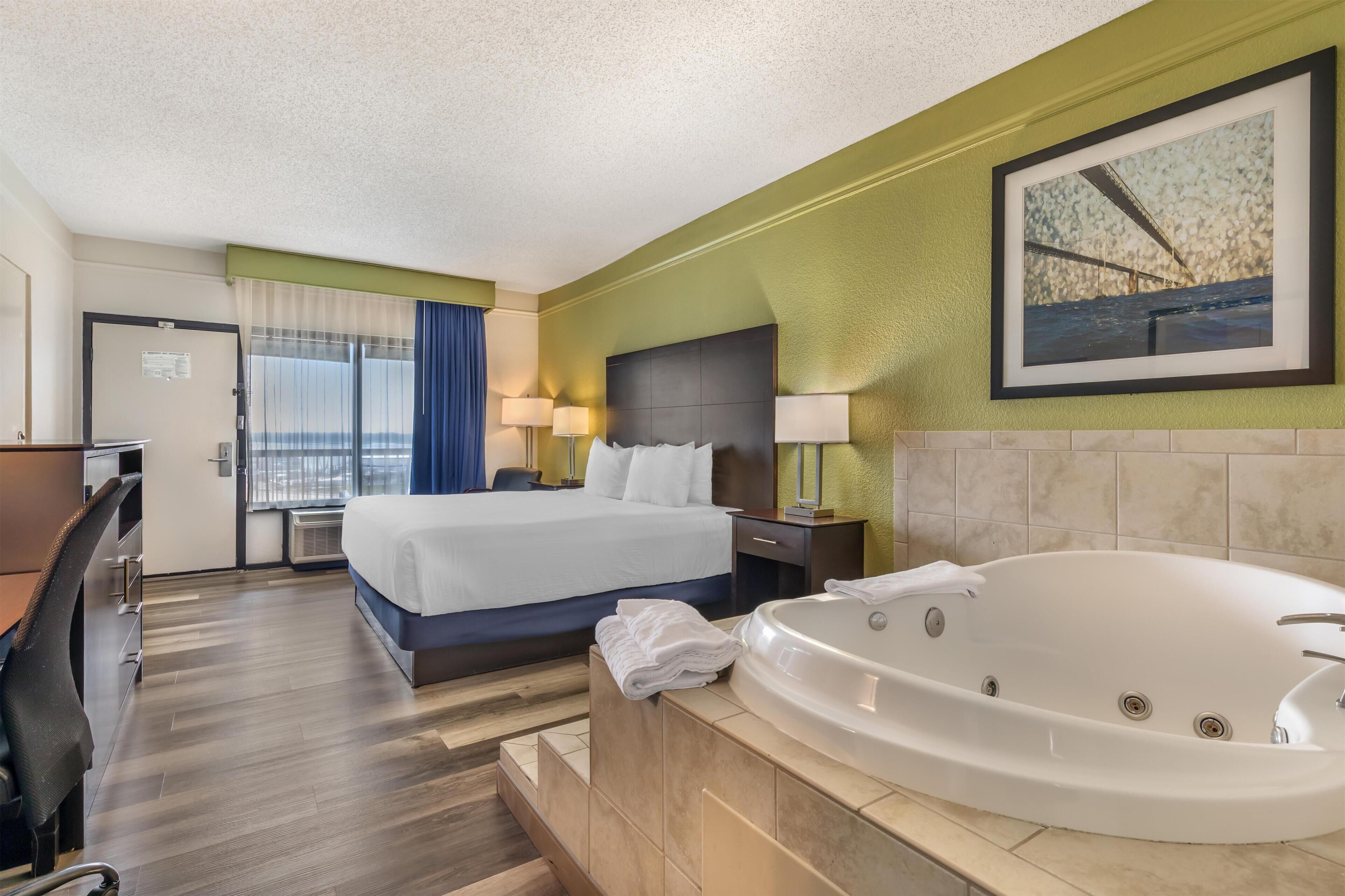 A hotel room with a bed, a desk, a chair, a large window with curtains, a wall-mounted painting, and a bathtub.