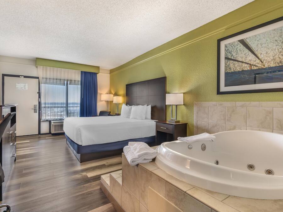 A hotel room with a bed, a desk, a chair, a large window with curtains, a wall-mounted painting, and a bathtub.