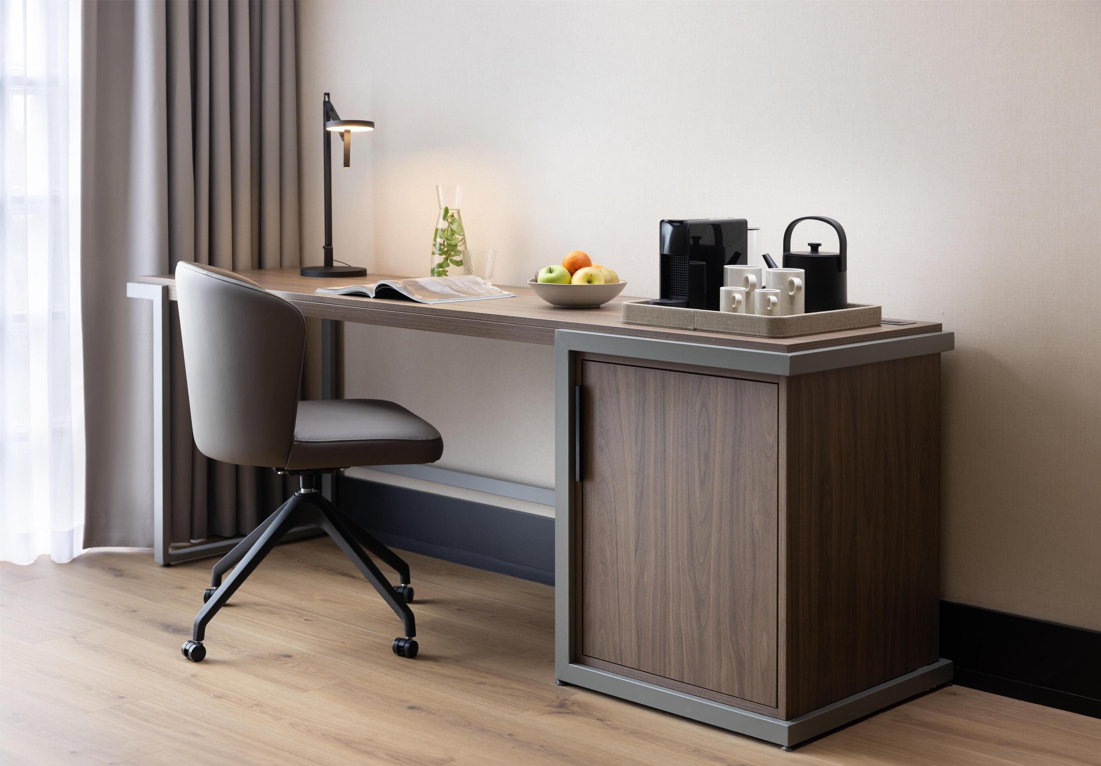 A modern office desk with a chair, a coffee maker, a kettle, and a bowl of fruit.