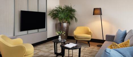 Ein modernes Wohnzimmer mit einem Flachbildfernseher, zwei Sesseln, einer Couch, einem Couchtisch, einer Stehlampe und einer Zimmerpflanze.