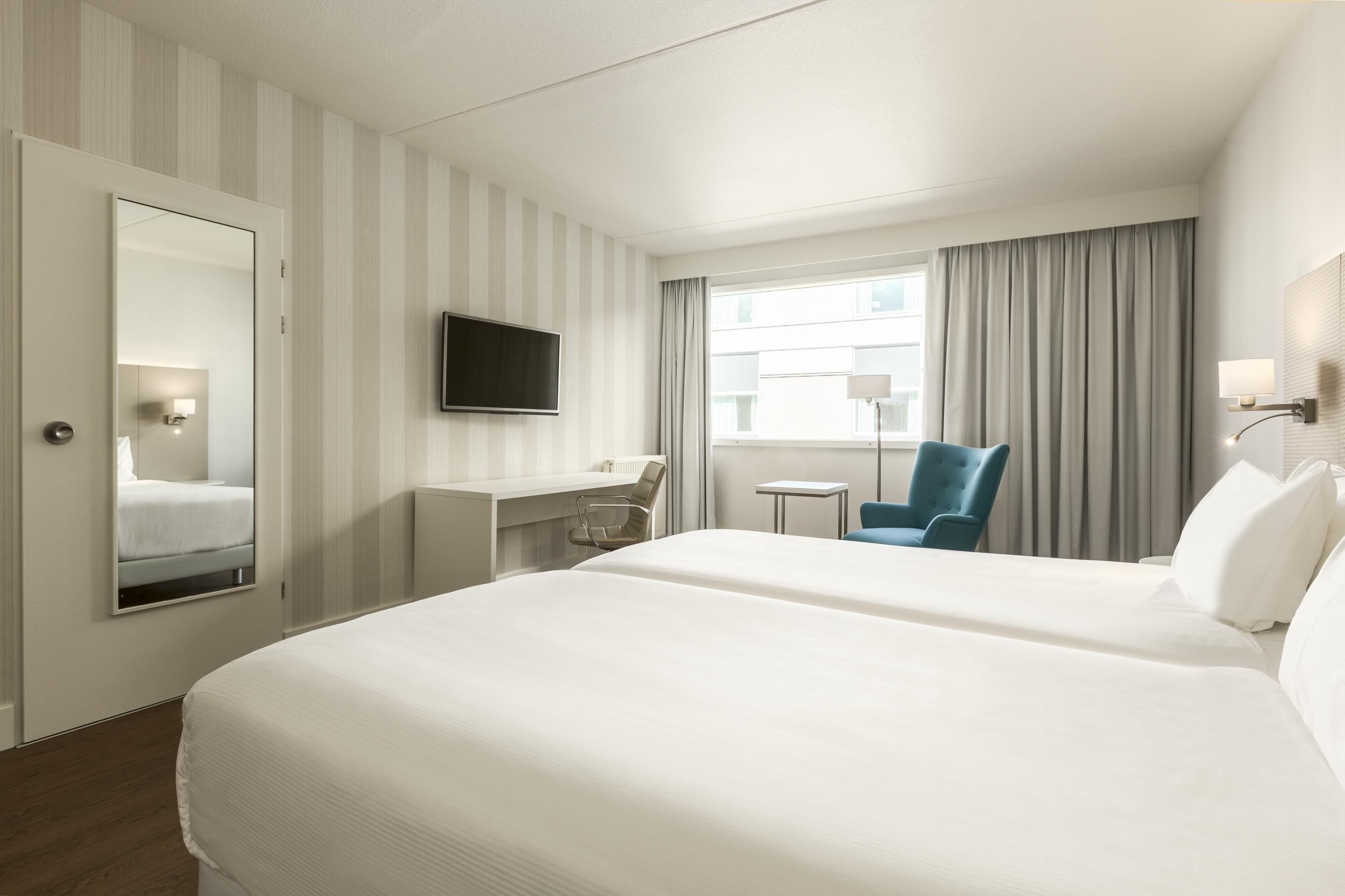 A hotel room with a large bed, a desk with a chair, a TV mounted on the wall, and a window with curtains.