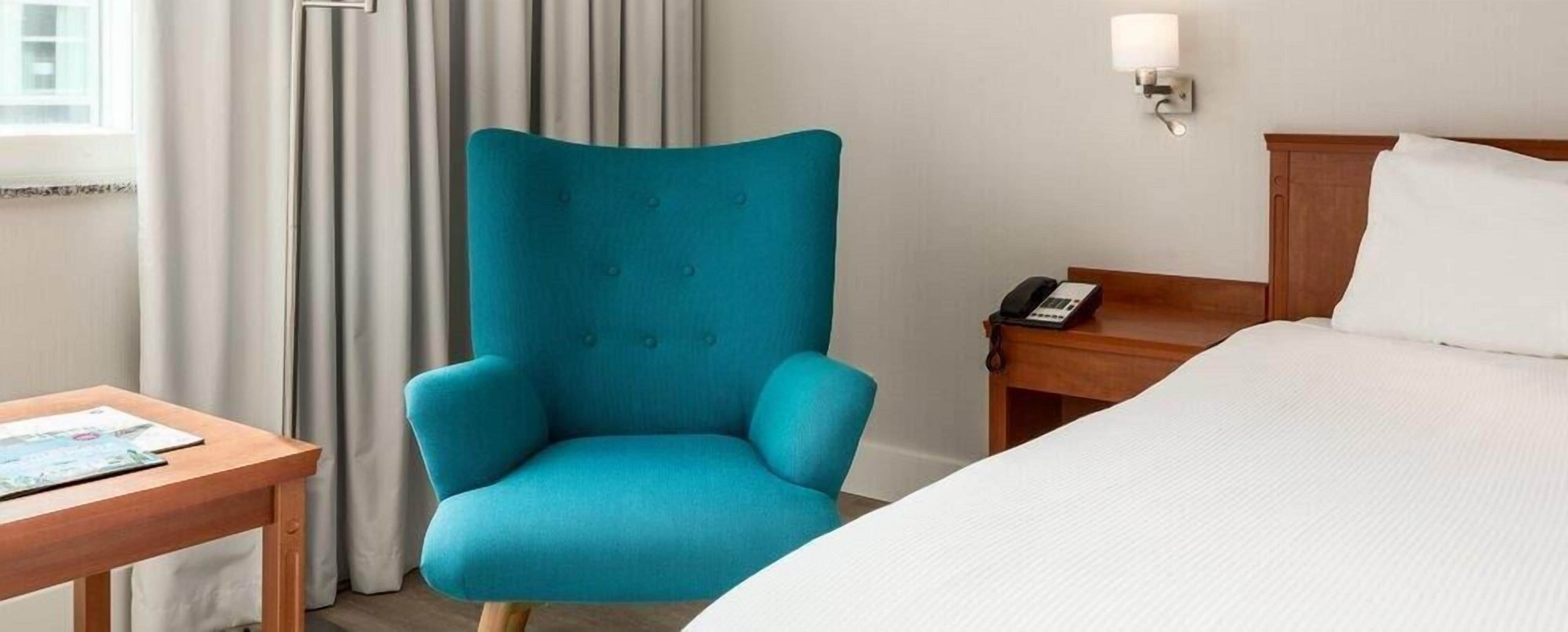 Una habitación de hotel con un sillón verde azulado, una mesita de noche de madera con un teléfono, una cama con ropa de cama blanca y una ventana con cortinas.