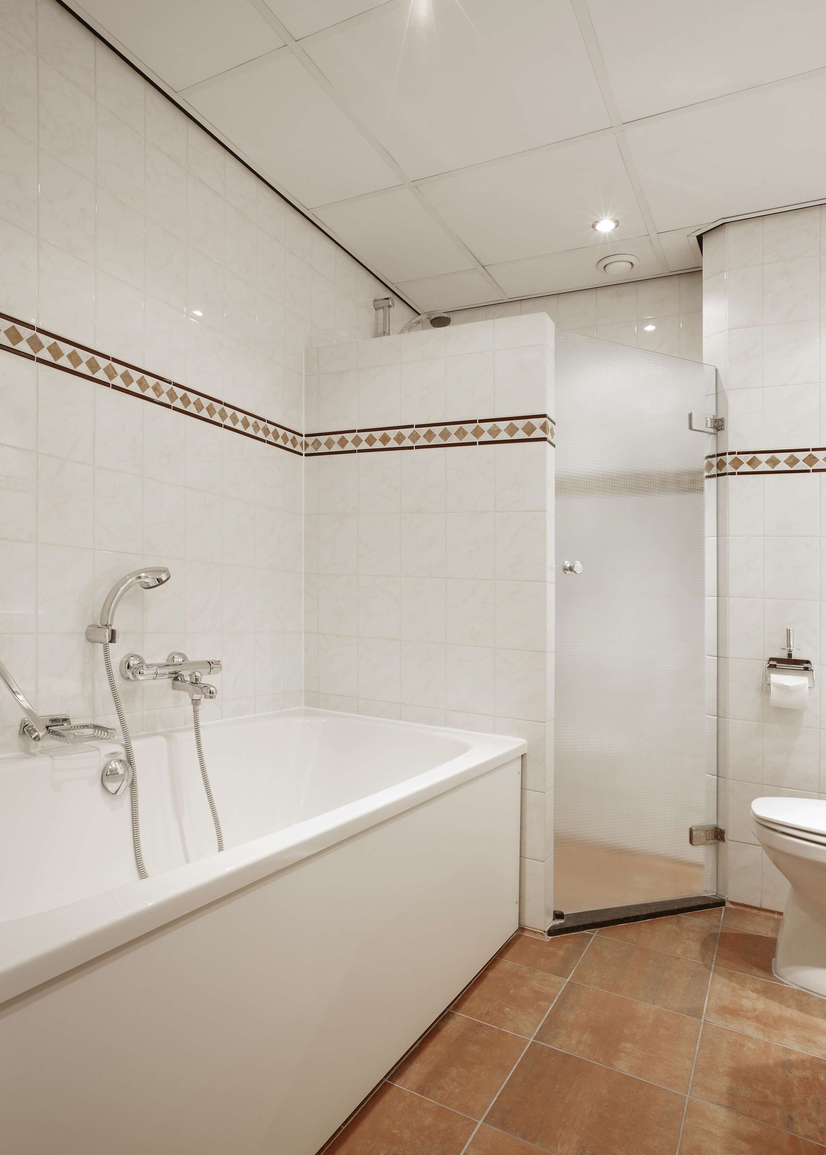 A bathroom with a white bathtub, a shower, and a toilet. The walls are tiled with a white and brown pattern.