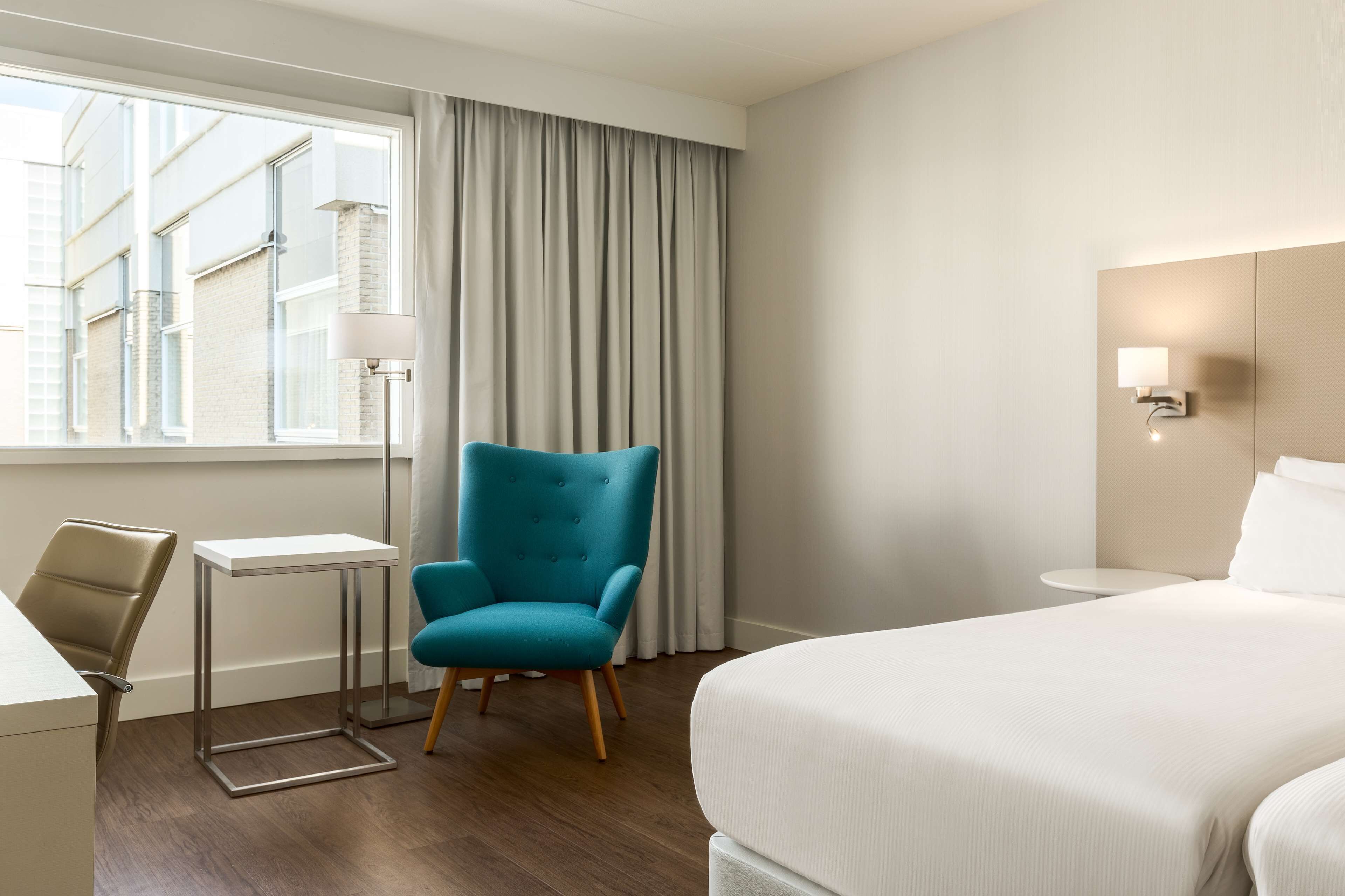 Ein Hotelzimmer mit Bett, Schreibtisch, Stuhl, Lampe und Blick auf Gebäude durch das Fenster.