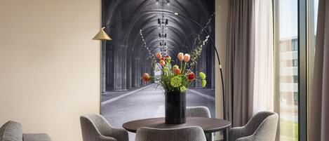 A modern hotel room with a round dining table, grey upholstered chairs, a vase with flowers, and a large framed picture of a cathedral.