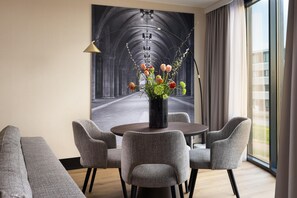 Ein modernes Hotelzimmer mit einem runden Esstisch, grauen Polsterstühlen, einer Blumenvase und einem großen gerahmten Bild einer Kathedrale.