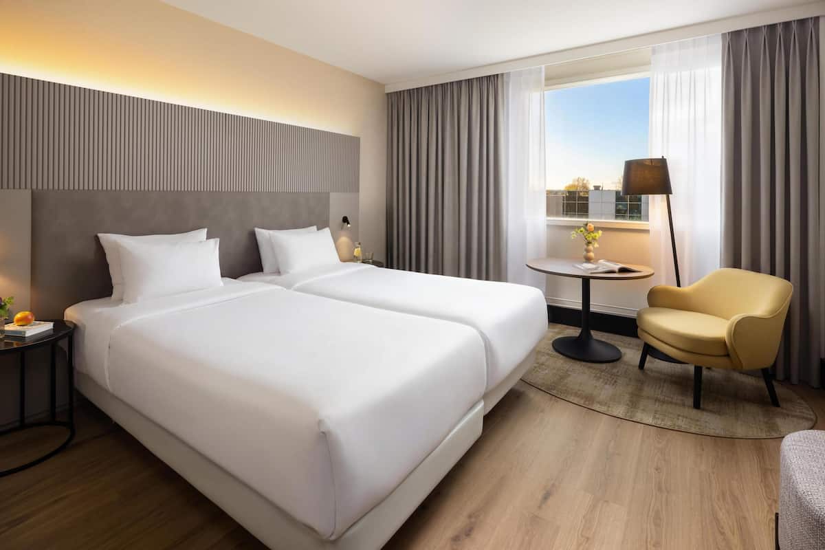 Habitación de hotel con dos camas, una mesita redonda pequeña, una silla y una mesita de noche con lámpara.