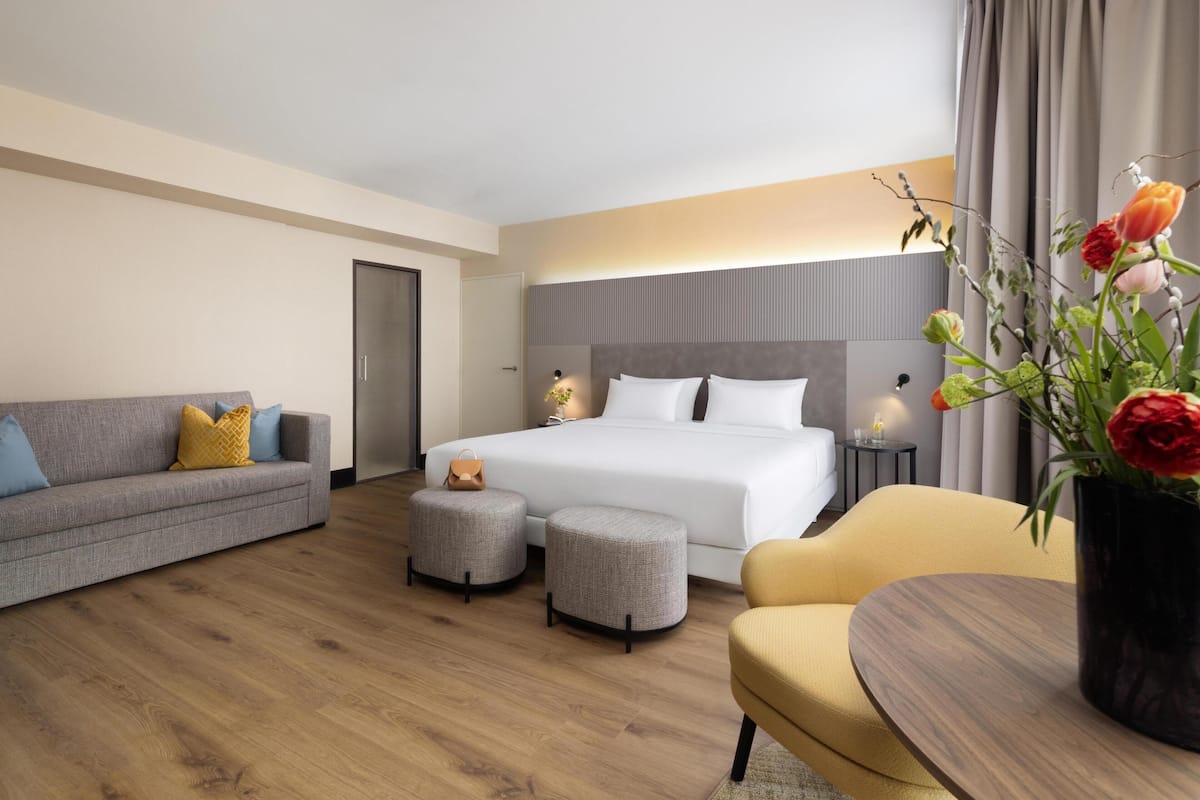 Una habitación de hotel moderna con cama, sofá, reposapiés y una mesa con un jarrón de flores.