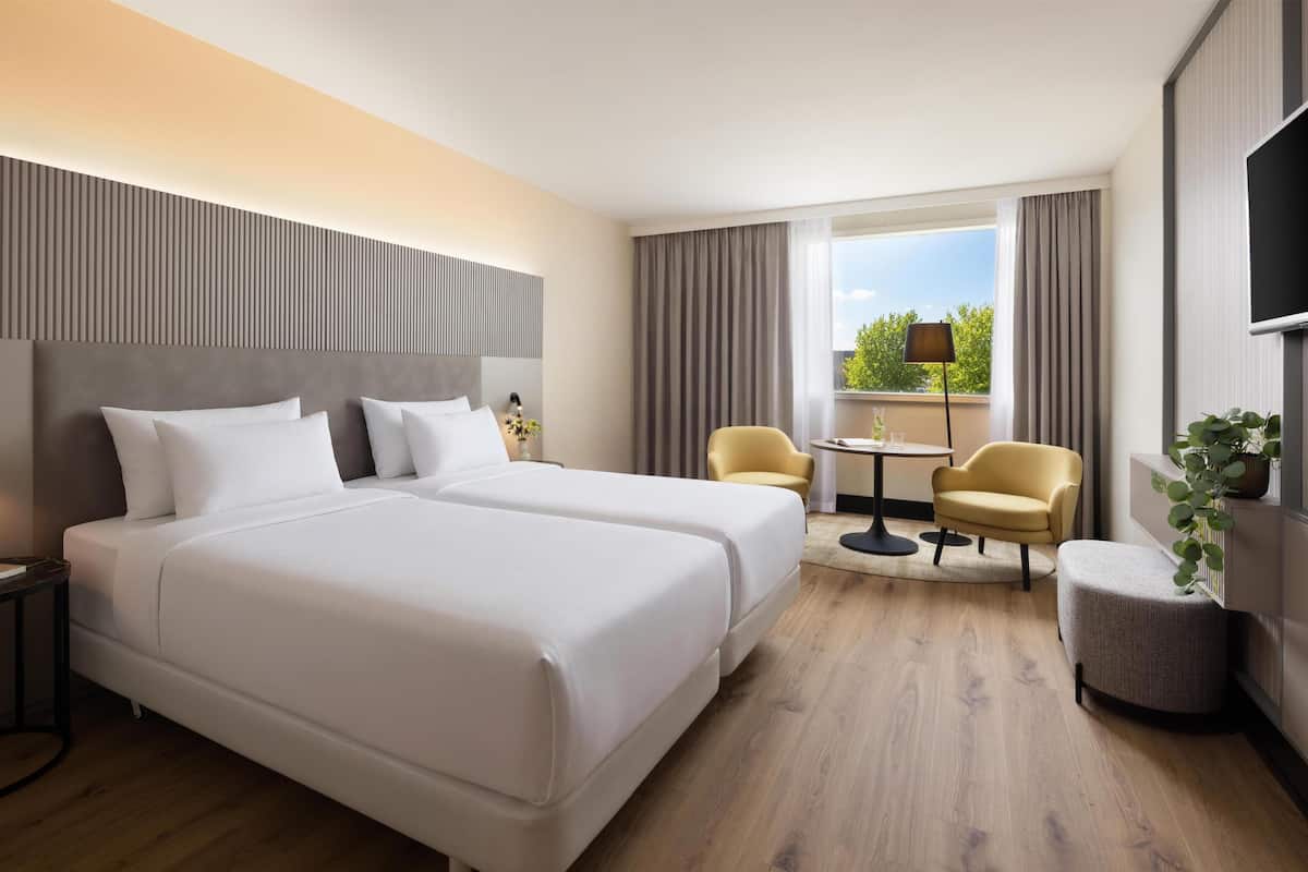 Habitación de hotel con dos camas, una mesita pequeña, dos sillas, un televisor y una ventana con cortinas.