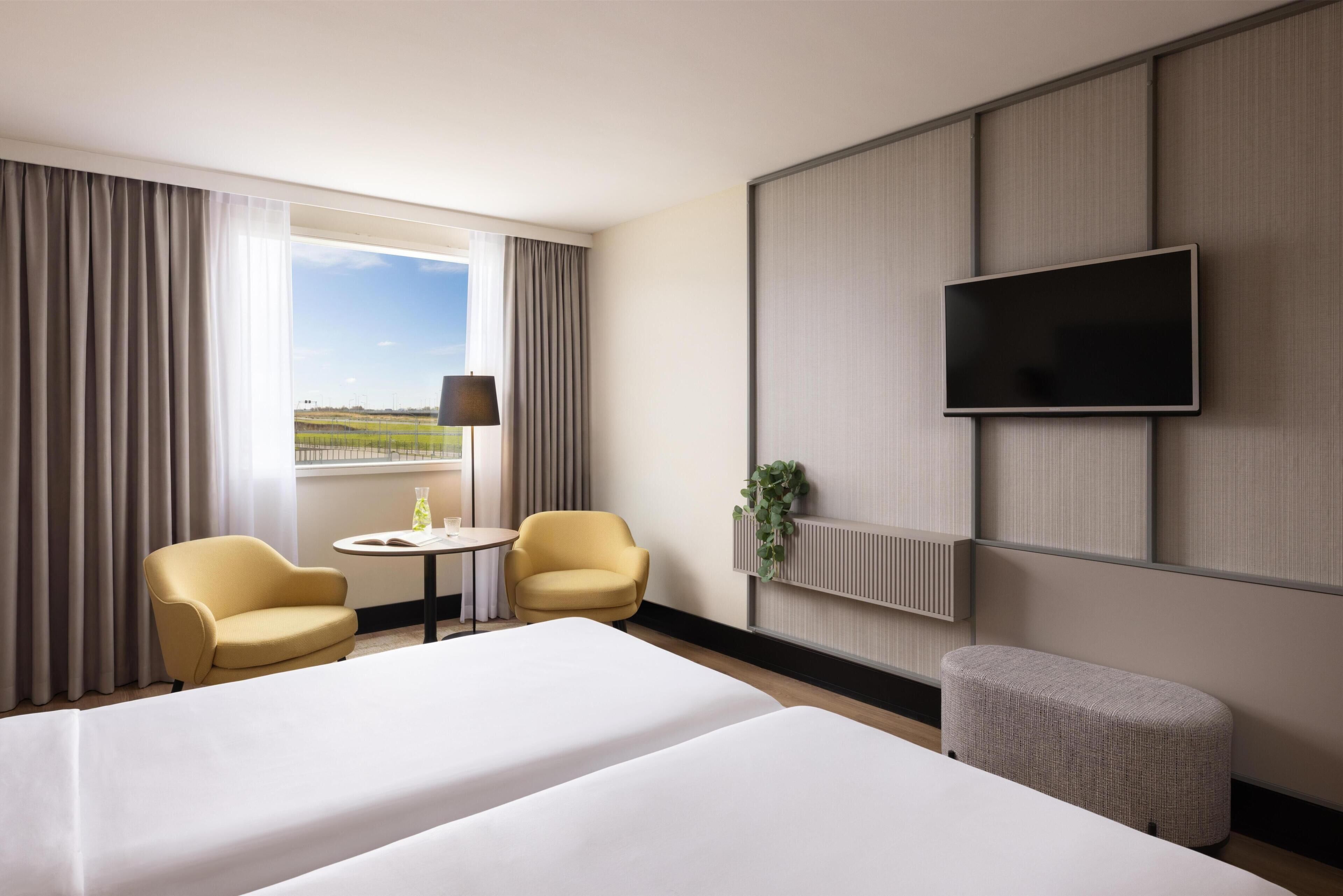 Habitación de hotel con una cama grande, dos sillones, una mesita, un televisor montado en la pared y una ventana con cortinas.
