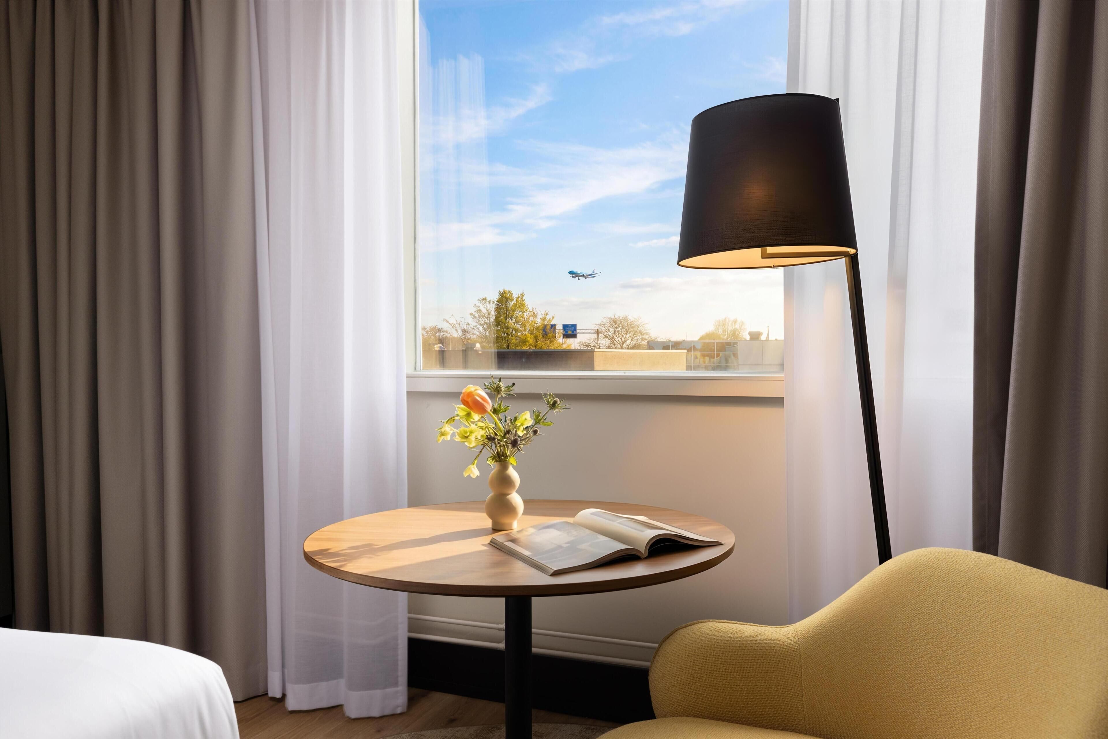 A hotel room with a bed, a round table with a book and a vase of flowers, a lamp, and a window with a view of the sky and a plane.